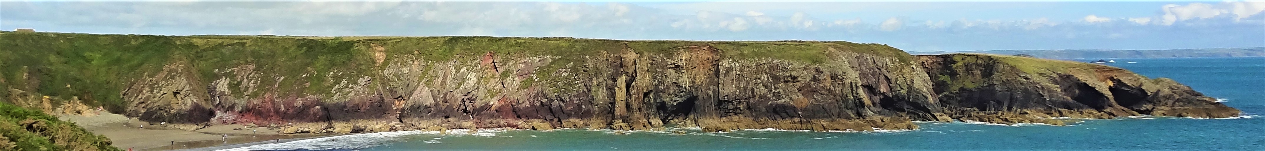 Panoramic view across a sunny bay from the cliffs on one side of it, looking at the cliffs on the other side, with a sandy beach on the left to a blue-green sea on the right. The cliffs are flat-topped, green-grey on the left, becoming maroon red in the middle, before turning yellow near the low tide mark, and eventually black in colour towards the headland.