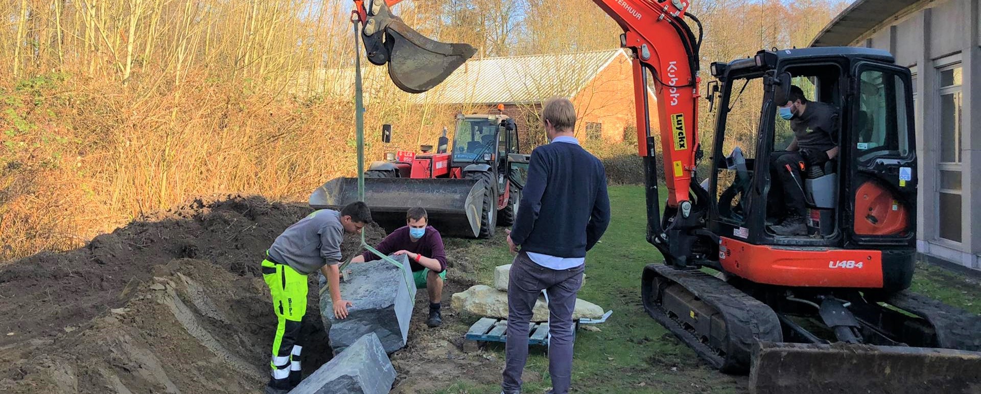 Three people are positioning a large block of grey stone at a 45 degree angle in a trench. One person is driving a digger from which the rock is suspended by a green strap. The other two people are standing and crouching by the block adjusting its angle. There is a fourth person, presumably supervising, standing between the digger and the trench, watching proceedings.