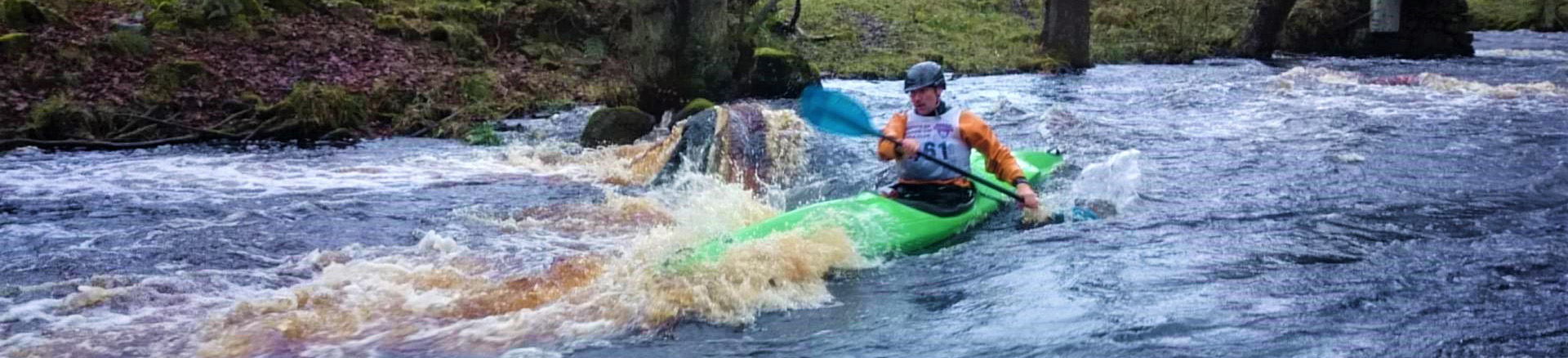 A kayaker in a yellow top and long green boat races past a rock on river-right of a Grade 2 white water river.